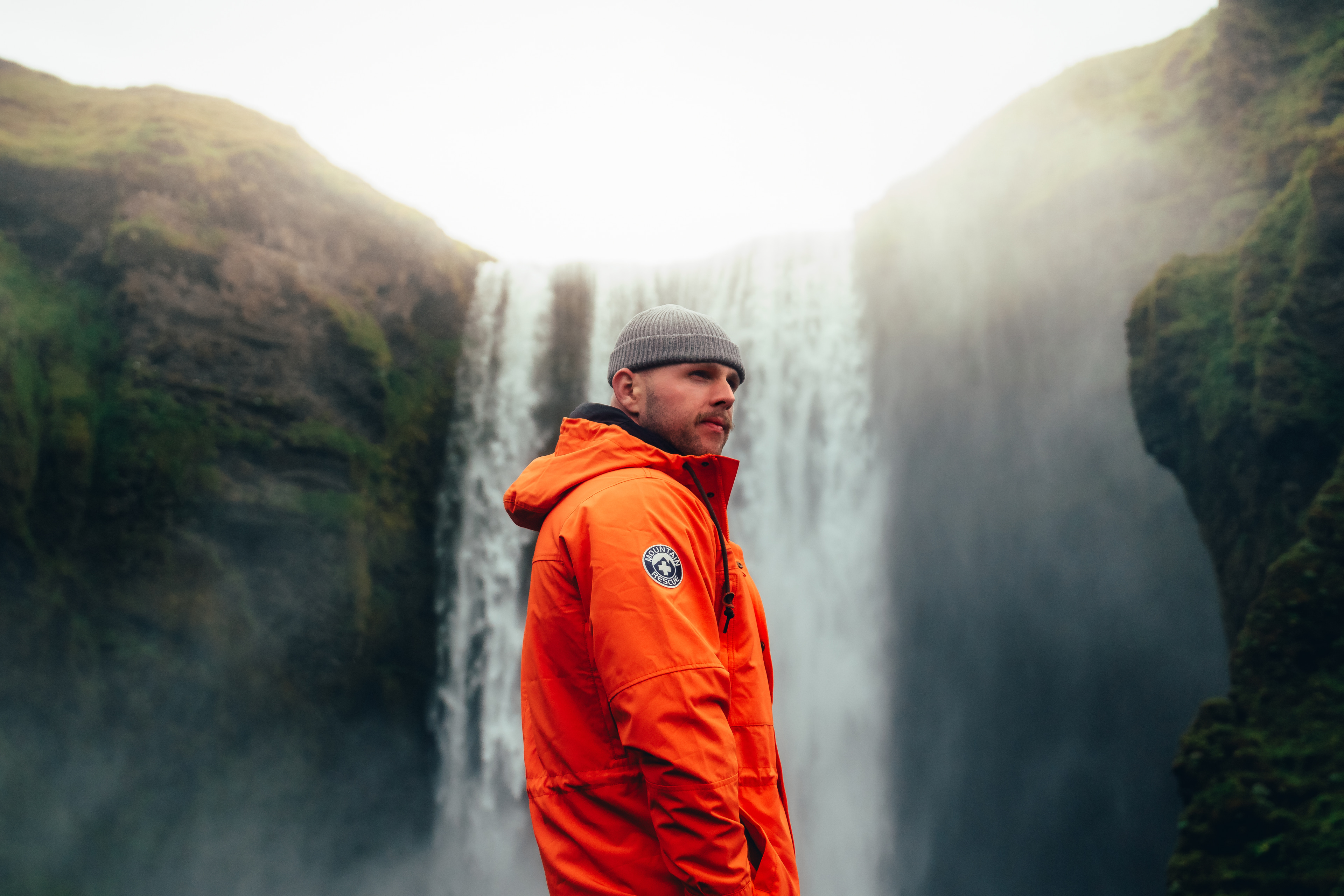 Max Loew in an orange Alpine Design shell jacket and beanie before a misty Icelandic waterfall