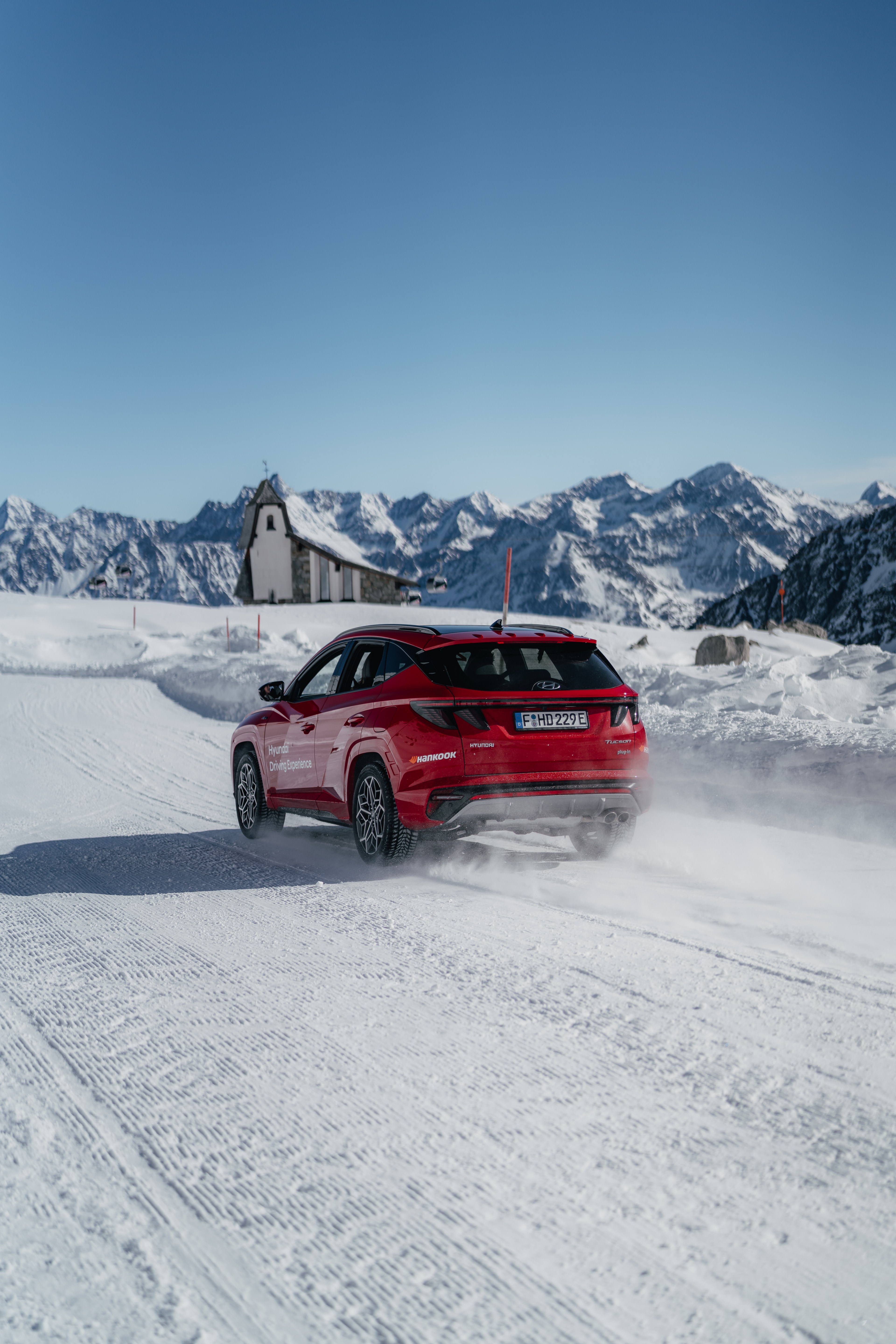 Red Hyundai Tucson on an alpine snow road, a mountain chapel and snowy peaks behind