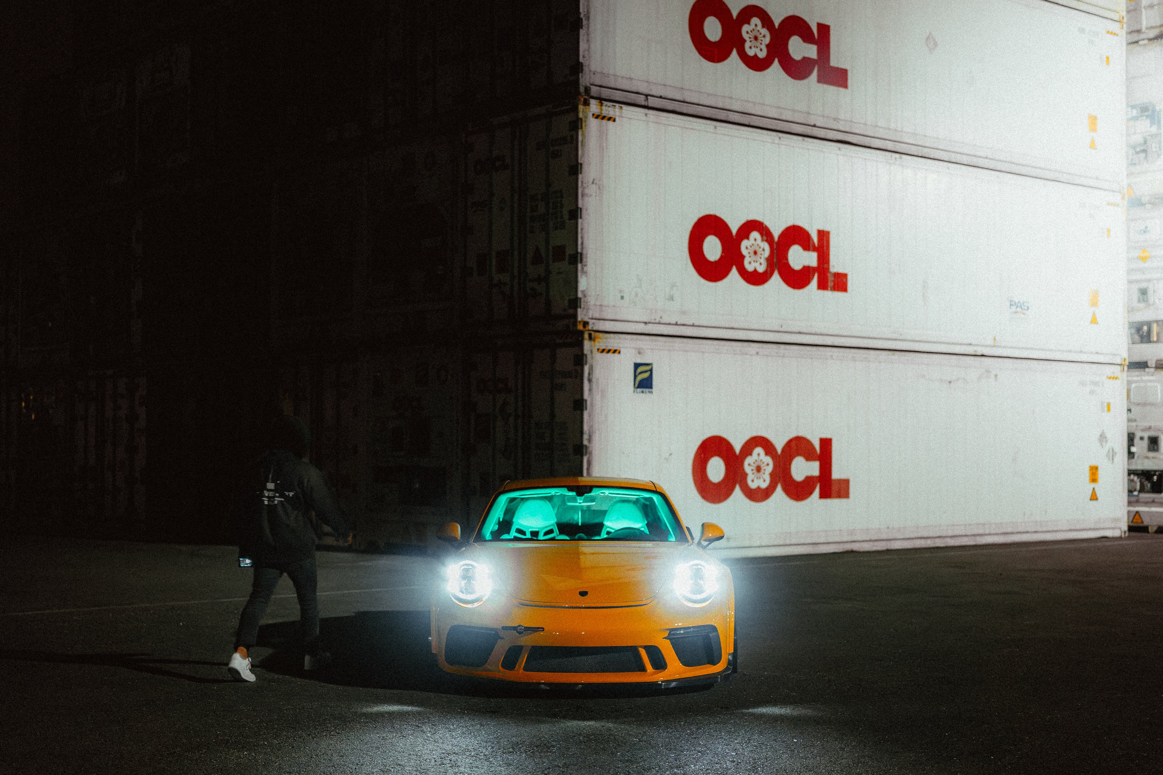 Yellow Porsche 911 GT3 at night against OOCL shipping containers, headlights blazing, a figure walking toward it