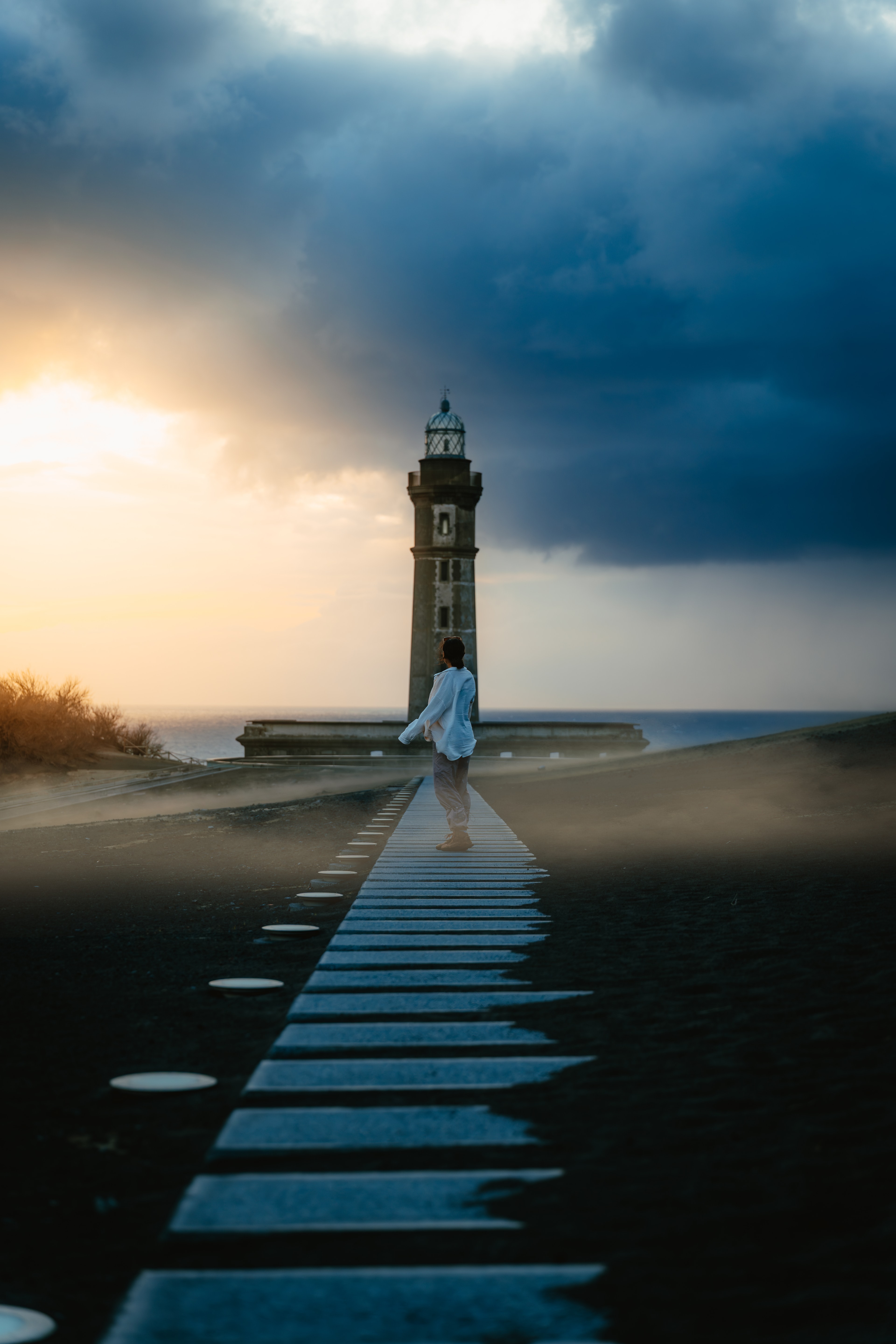 A lighthouse at the end of a long stone breakwater under a stormy sunset