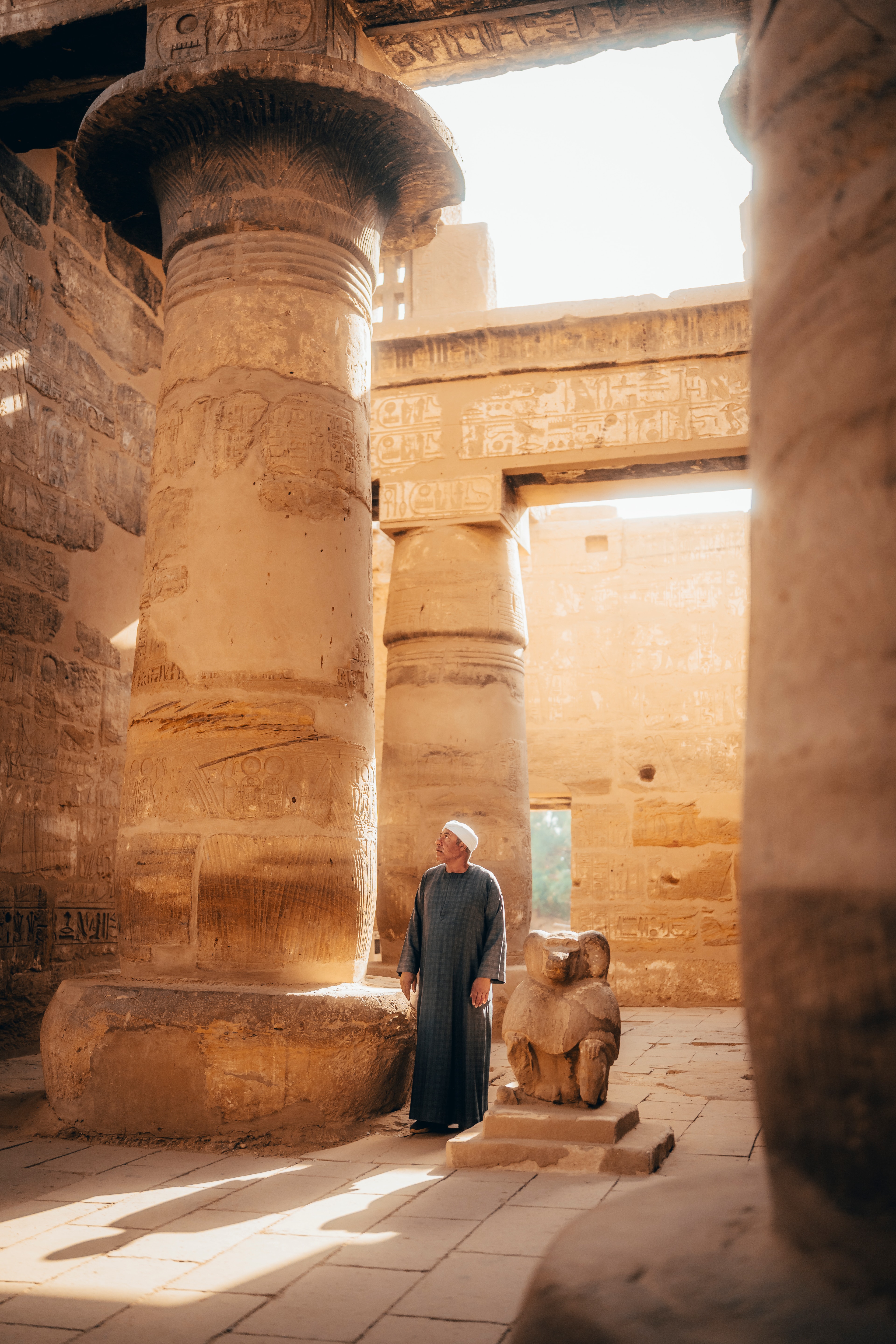 A temple guardian in a blue robe beside a small sphinx statue, dappled light between the columns