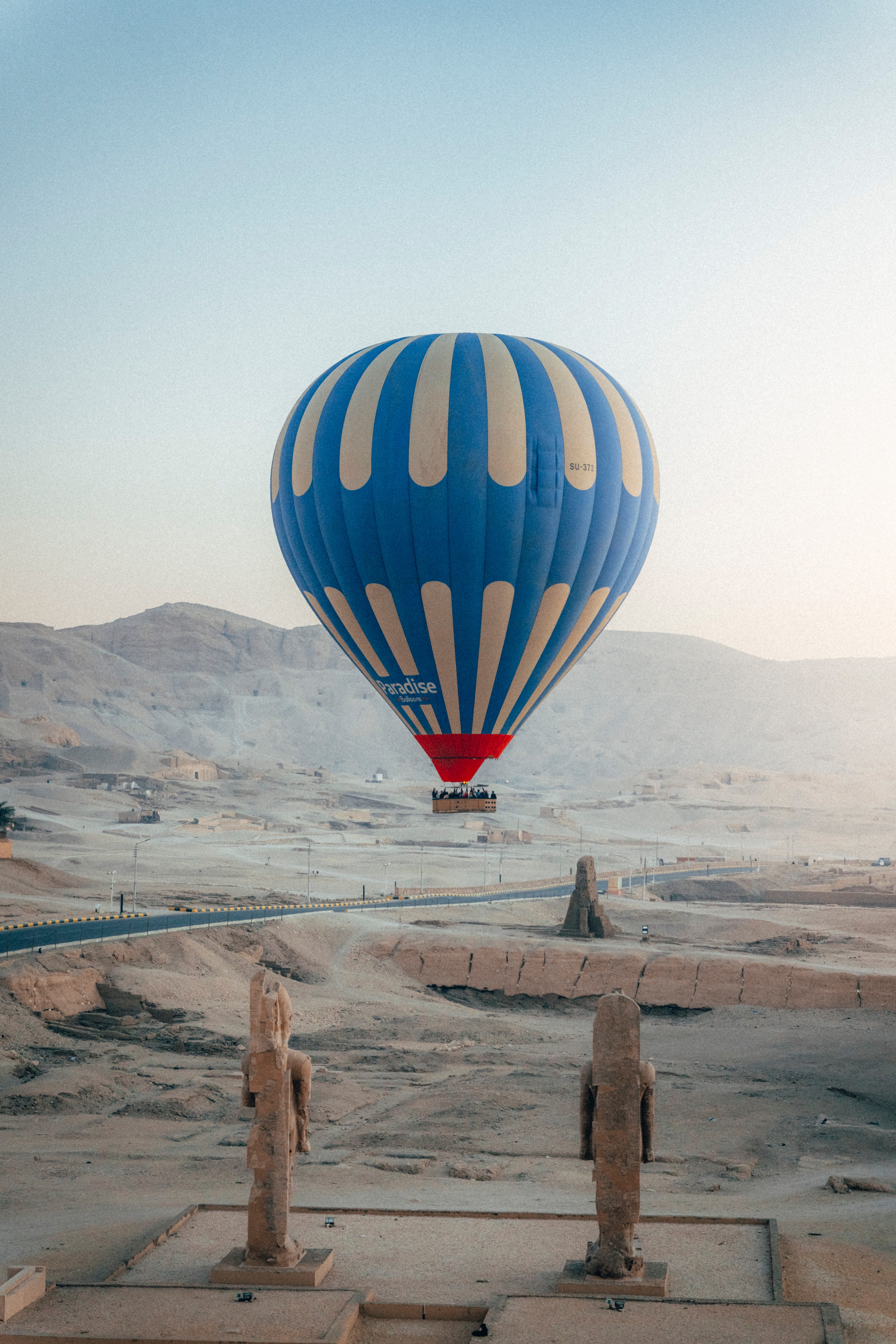A blue-and-cream hot air balloon above the Colossi of Memnon at first light