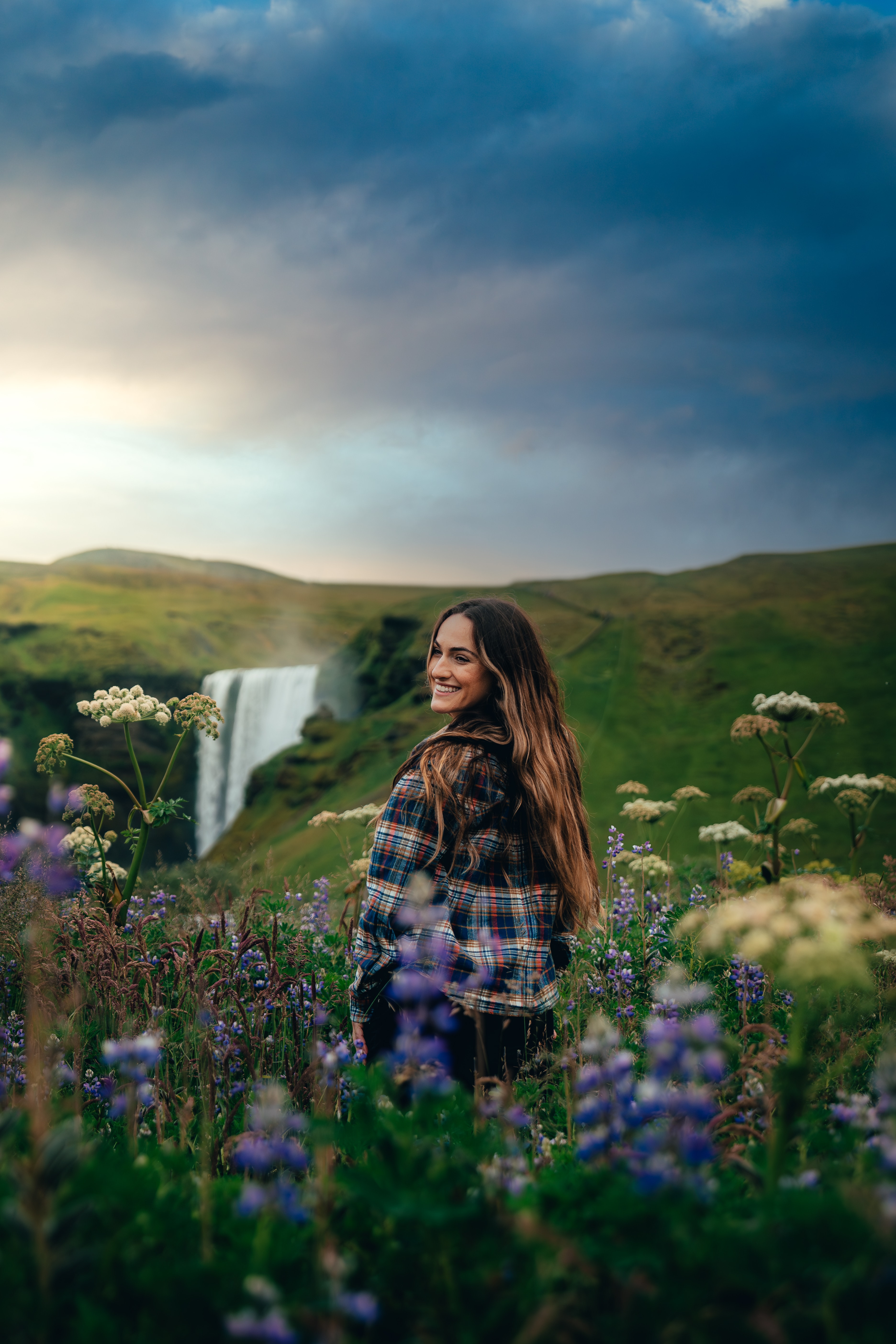 A woman in flannel in a field of lupines, Skogafoss and a low sun behind her