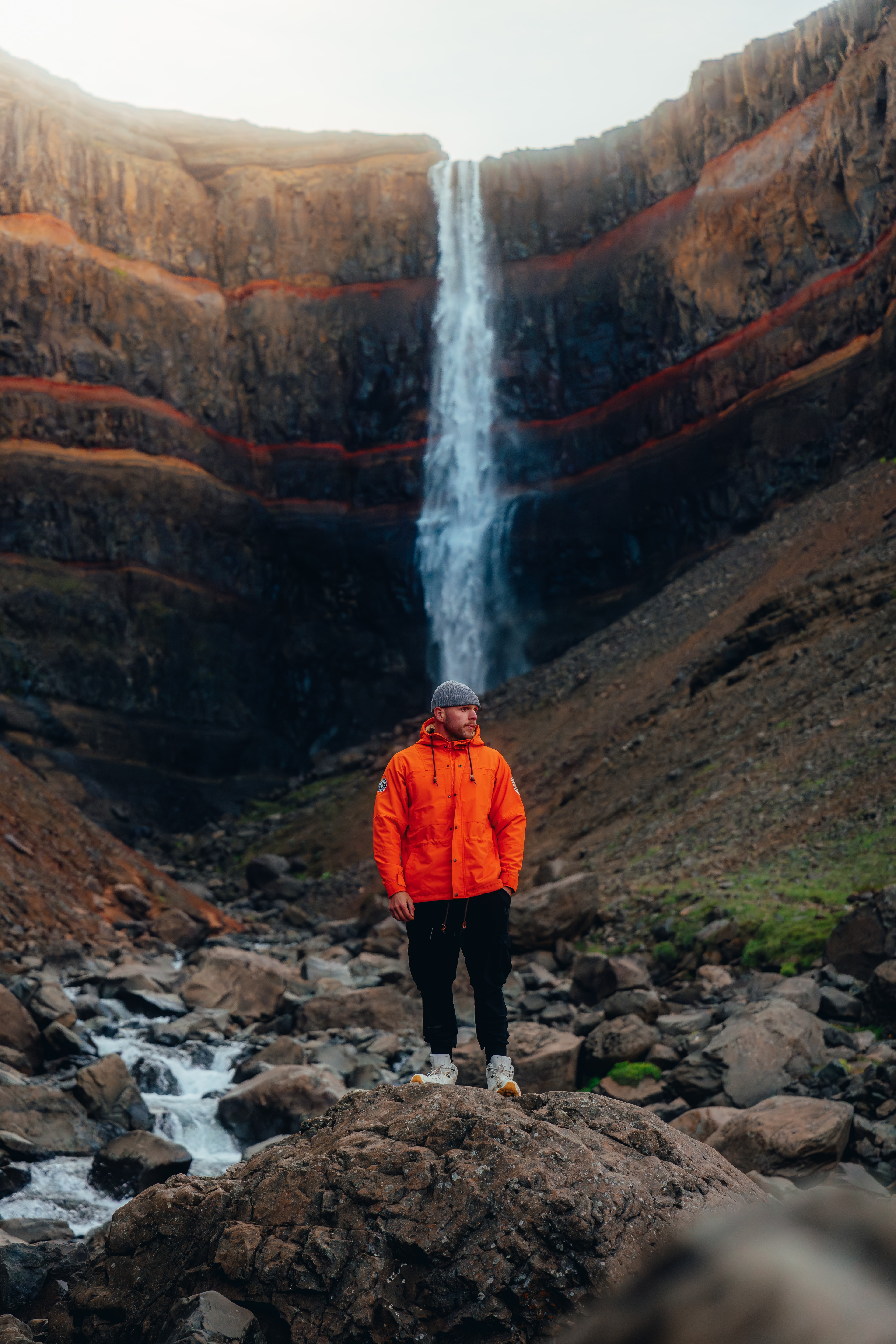 A figure in an orange shell beneath the red-banded cliffs of Hengifoss