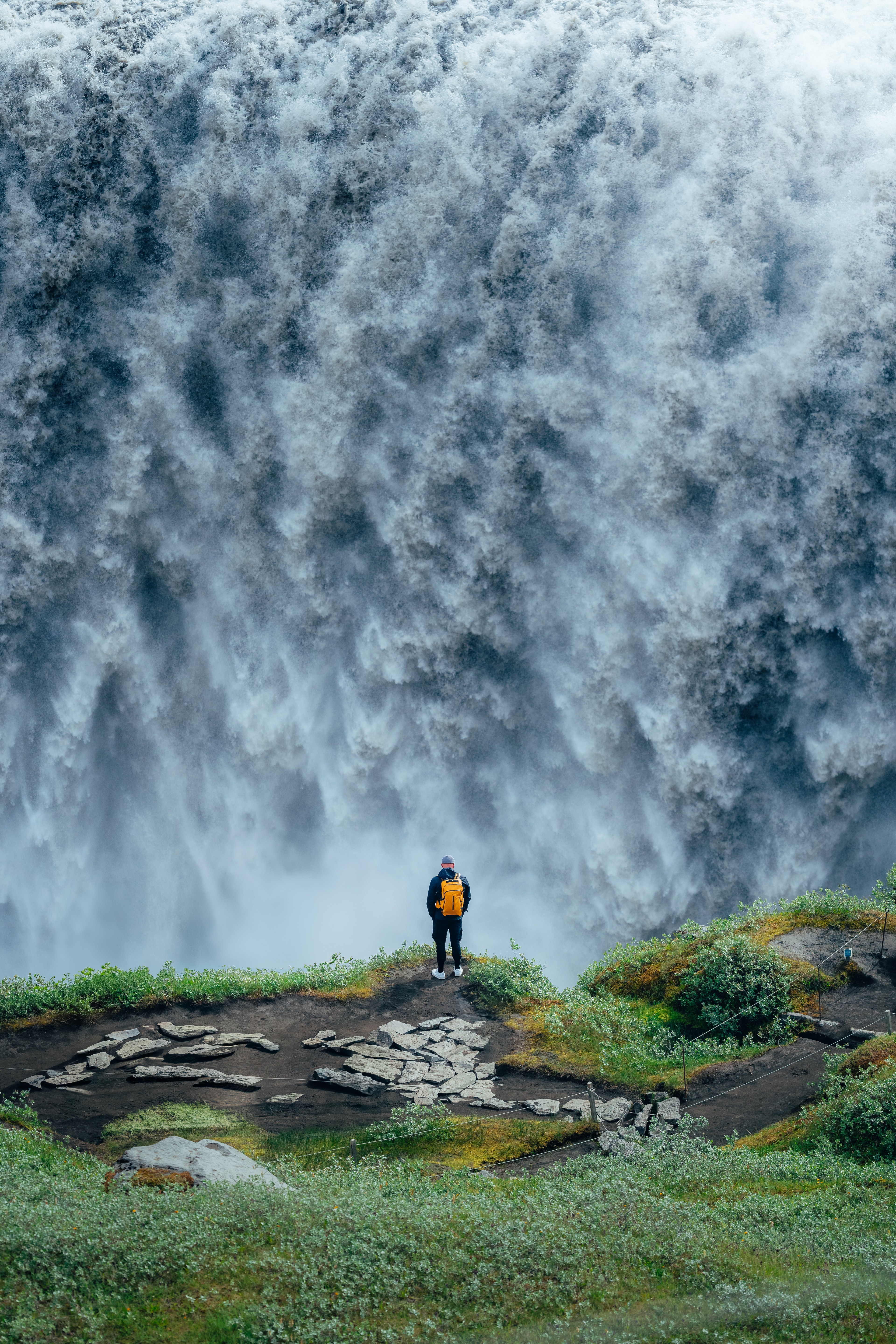 A tiny figure on a green ledge before the immense white wall of Dettifoss